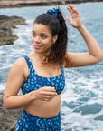 Model standing on a rocky coastline with waves crashing in the background, wearing NAUTRA’s Whale Shark Print Scrunchie. Inspired by the unique patterns of whale sharks, this ocean-conscious accessory ties together style and sustainability.