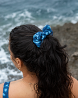 Model standing on a rocky coastline with waves crashing in the background, wearing NAUTRA’s Whale Shark Print Scrunchie. Inspired by the unique patterns of whale sharks, this ocean-conscious accessory ties together style and sustainability.