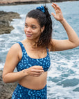 Model standing on a rocky coastline with waves crashing in the background, wearing NAUTRA’s Whale Shark Print Scrunchie. Inspired by the unique patterns of whale sharks, this ocean-conscious accessory ties together style and sustainability.