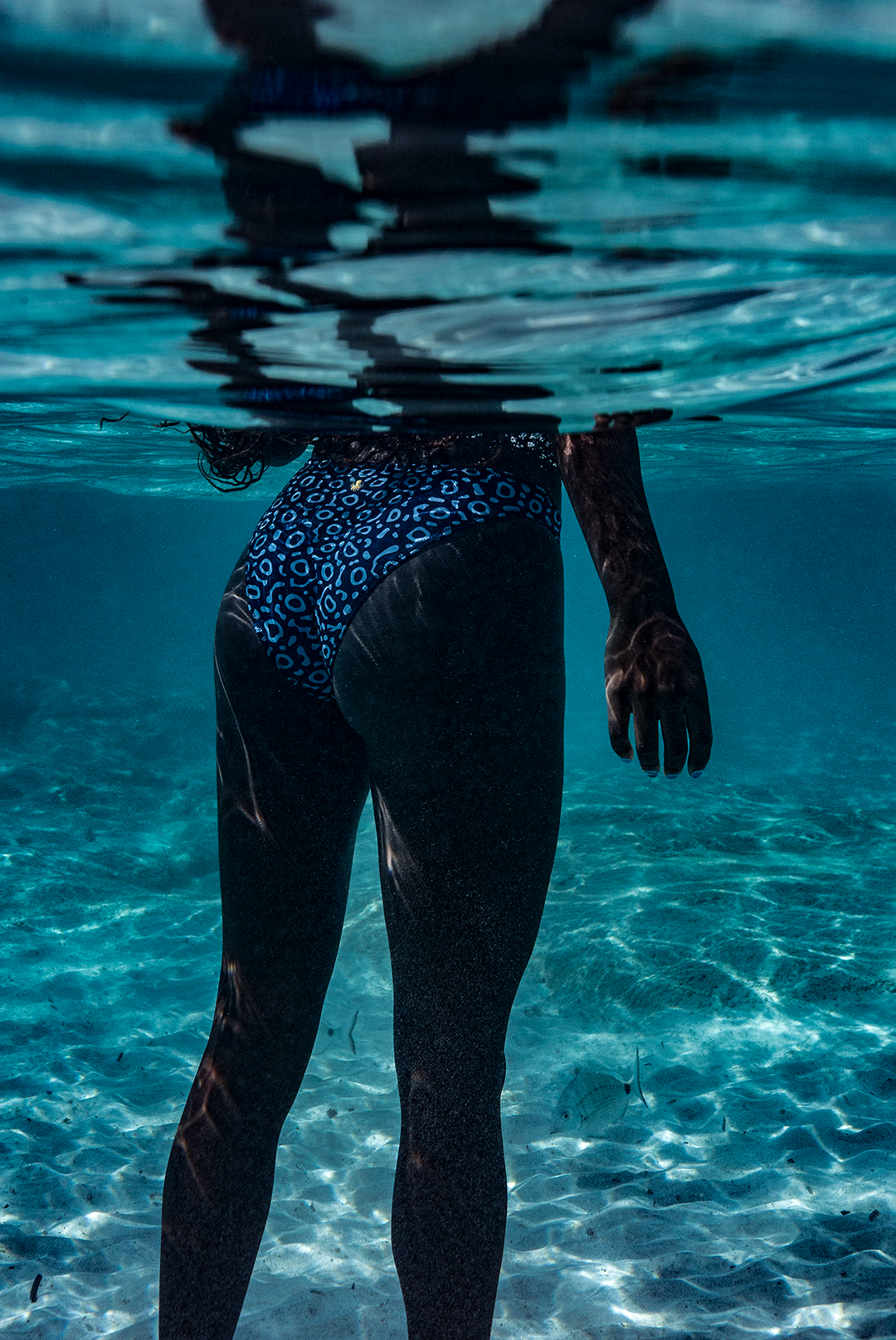 A woman wears NAUTRA’s Eagle Ray Print Bikini Bottoms on a sandy shore. These high-leg bikini bottoms (UK) are crafted from swimwear made from ghost nets, combining style and sustainability.

