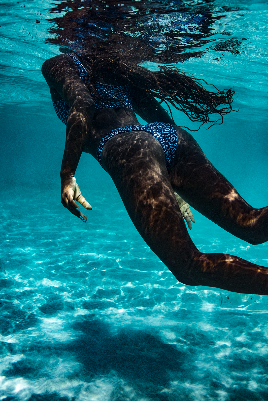 A woman wears NAUTRA’s Eagle Ray Print Bikini Bottoms on a sandy shore. These high-leg bikini bottoms (UK) are crafted from swimwear made from ghost nets, combining style and sustainability.
