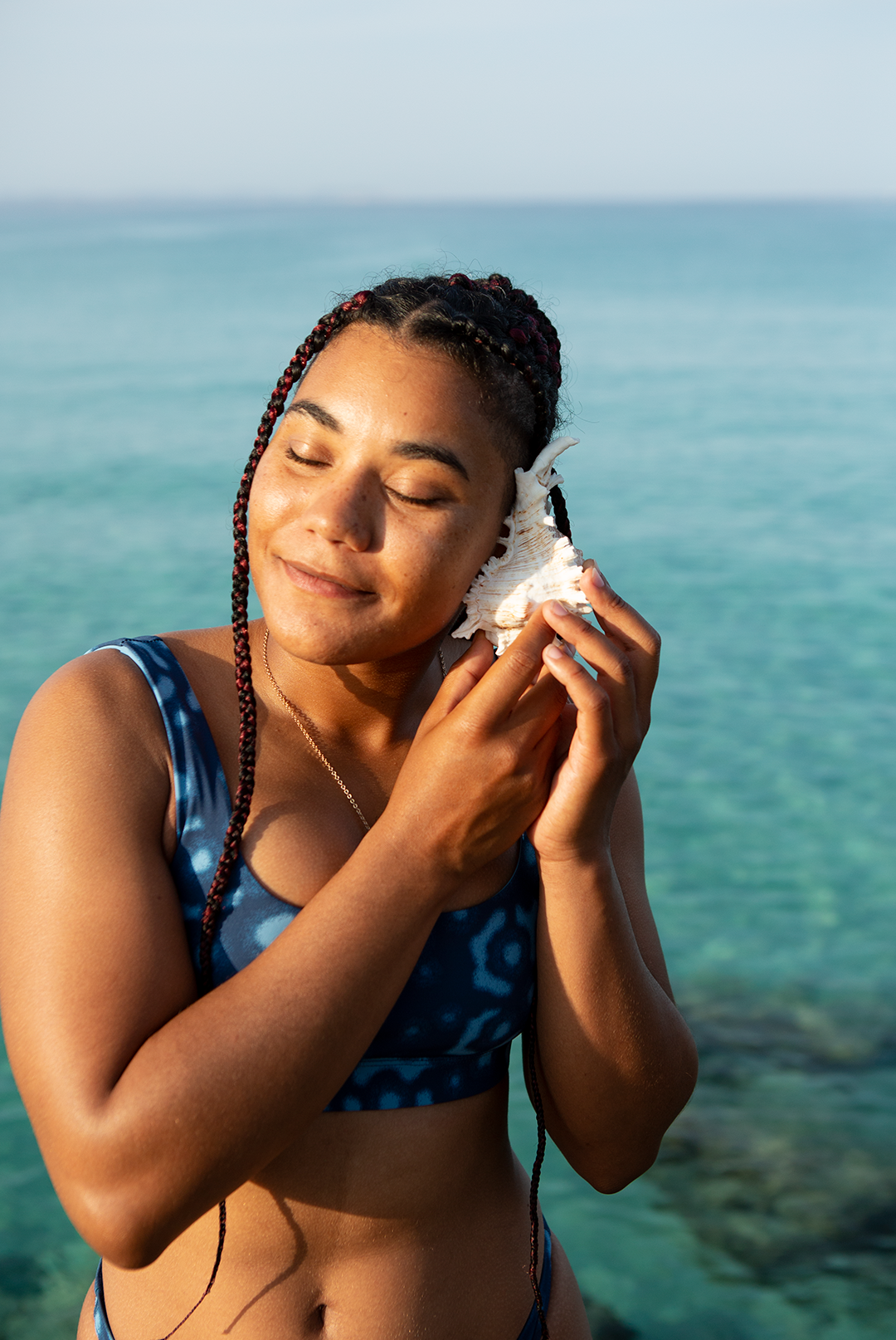 Girl on the beach wearing the whale shark print bikini top from NAUTRA listening in to a sea shell. Eco friendly swimwear UK.