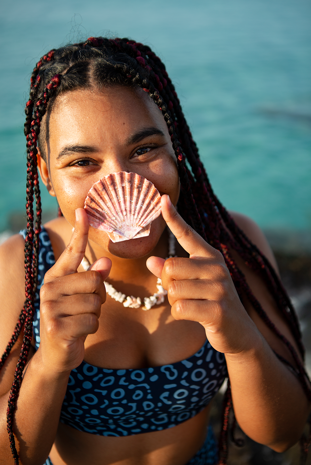 Girl smiling into the camera holding a shell on her nose. She is wearning NAUTRA sustainable swimwear UK. Eagle ray print scoop neck sports bra bikini top.