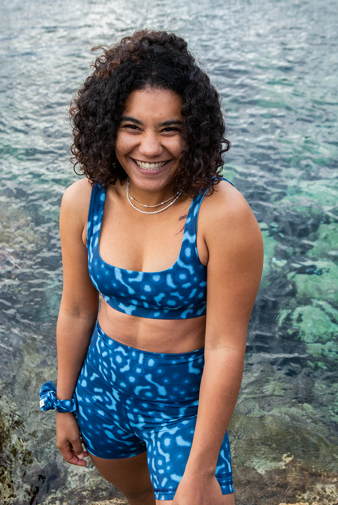 A woman wears NAUTRA’s Whale Shark Bikini Top on a rocky shore. This eco-friendly scoop neck sports bra is chlorine-resistant and made for land and sea.