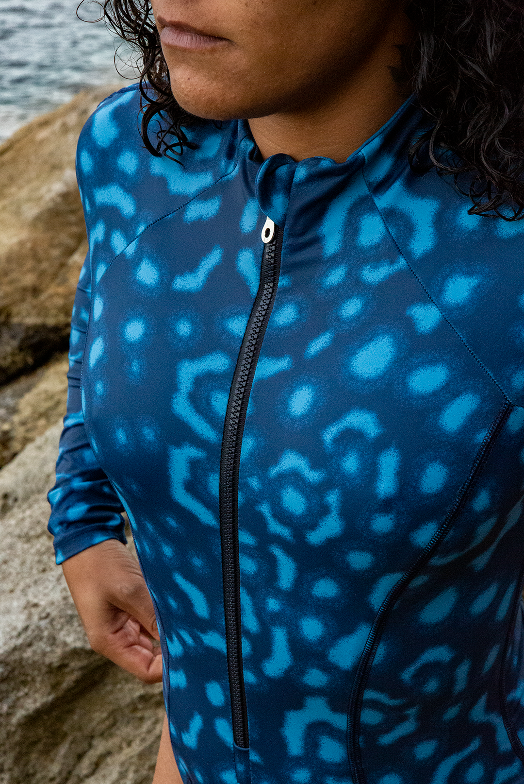A woman wears NAUTRA’s Women&#39;s Long-Sleeve One-Piece Swimsuit in Whale Shark Print on a rocky coast. This eco-friendly surf swimsuit is designed for adventure.