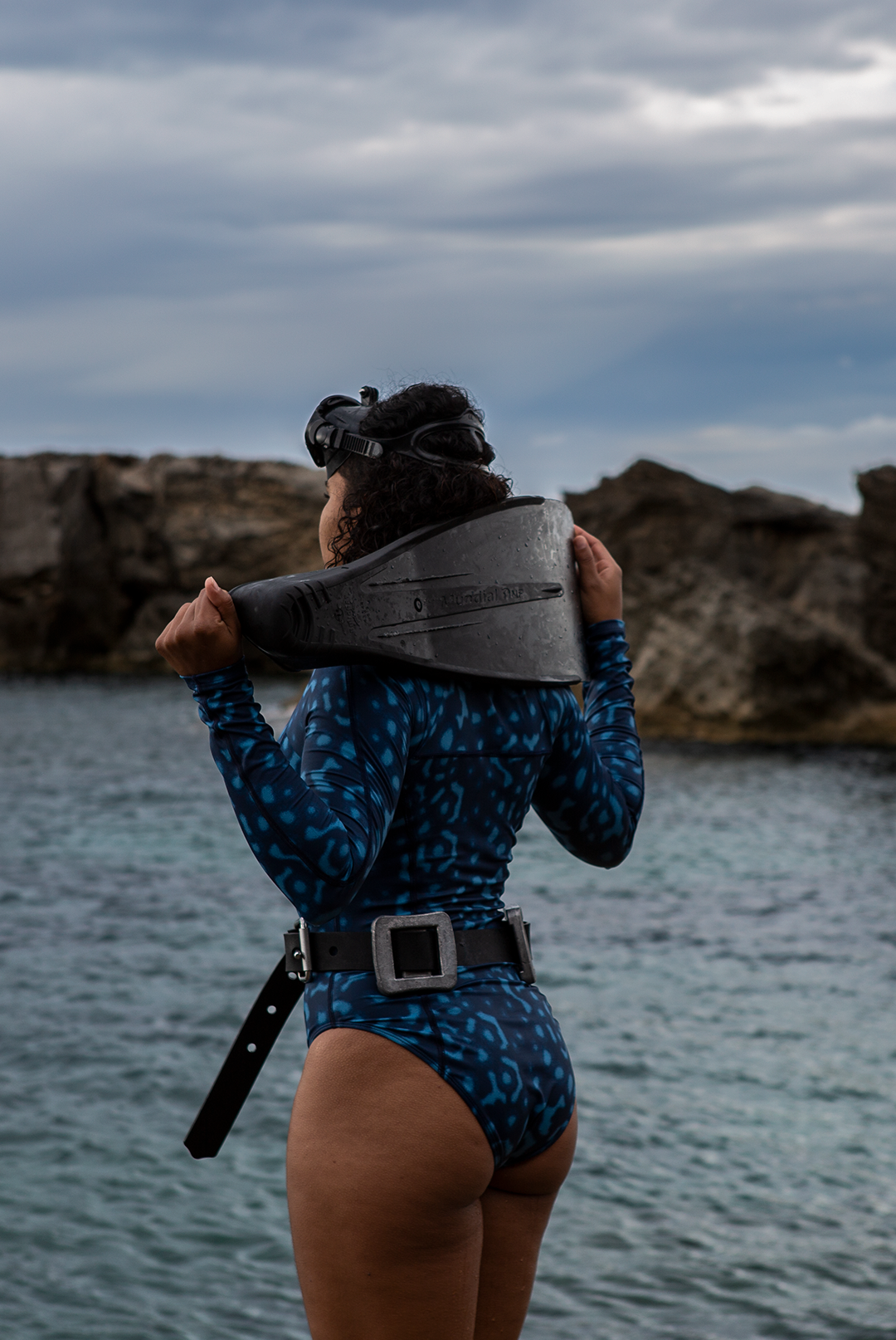 A woman wears NAUTRA’s Women&#39;s Long-Sleeve One-Piece Swimsuit in Whale Shark Print on a rocky coast. This eco-friendly surf swimsuit is designed for adventure.