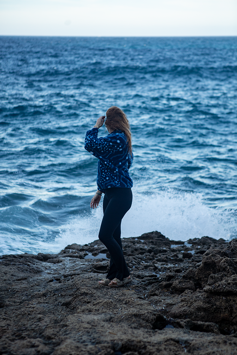 Model wearing the sustainable Whale Shark Fleece on rocky coastal cliffs with ocean waves crashing below.