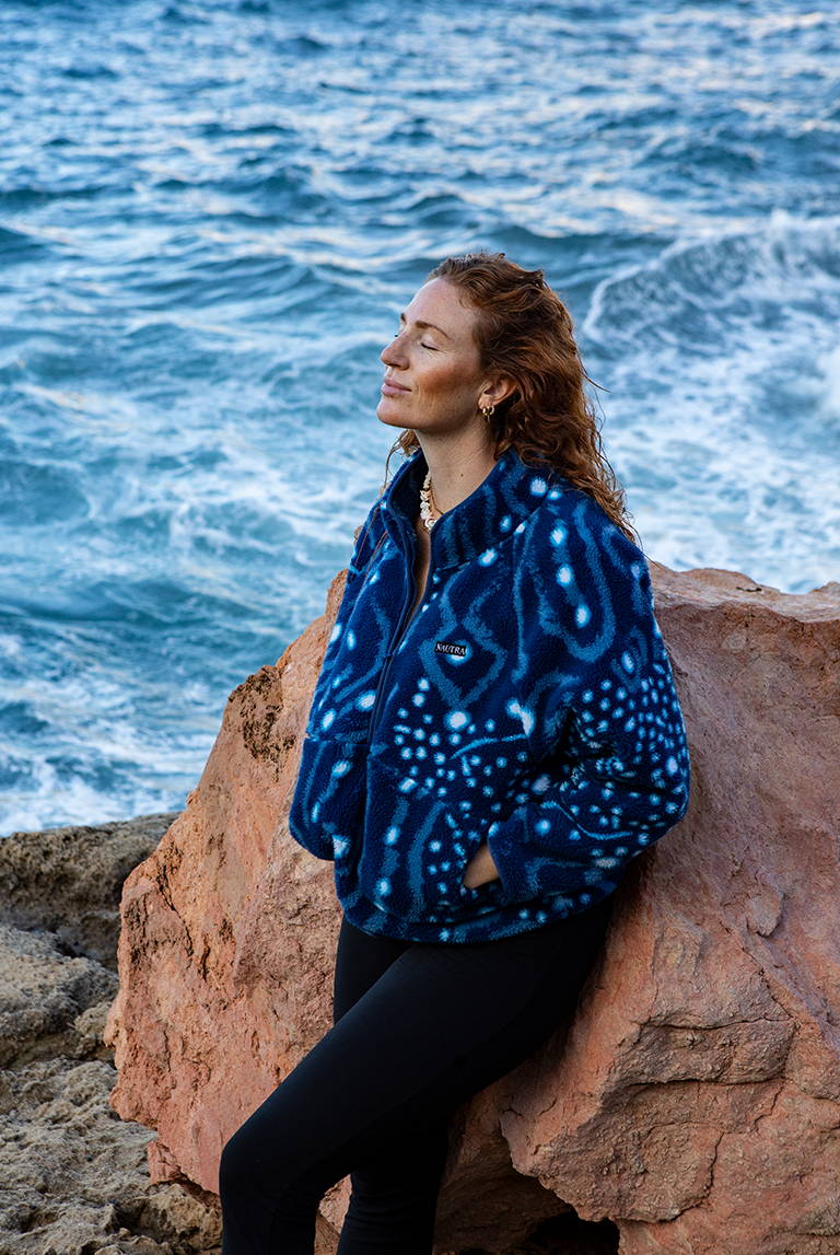 Model relaxing against a rock by the sea wearing the NAUTRA Whale Shark Fleece, a sustainable women’s fleece inspired by ocean giants.