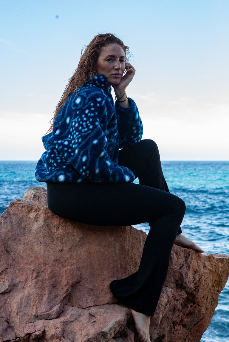 Woman sitting on coastal rocks wearing the Whale Shark Fleece by NAUTRA, made from recycled plastic bottles.