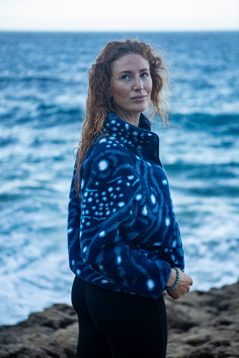 Woman wearing the NAUTRA Whale Shark Fleece made from recycled materials, standing by the ocean with waves in the background.