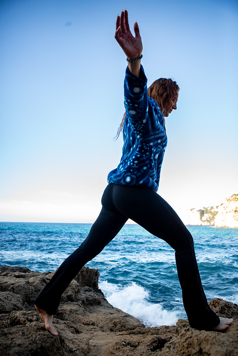 Model wearing the NAUTRA Whale Shark Fleece while stepping across coastal rocks by the sea. Ocean-inspired women’s recycled fleece designed for adventure.