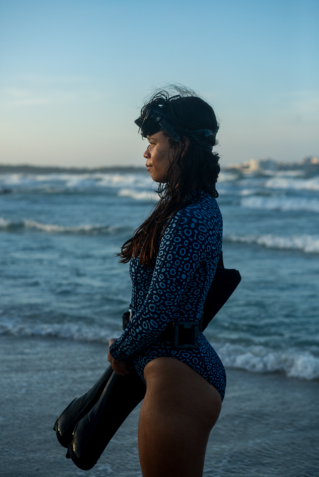 A woman wears NAUTRA’s Women's Long-Sleeve One-Piece Swimsuit in Eagle Ray Print on a sandy beach, with waves crashing behind her. This eco-friendly surf swimsuit is designed for adventure.