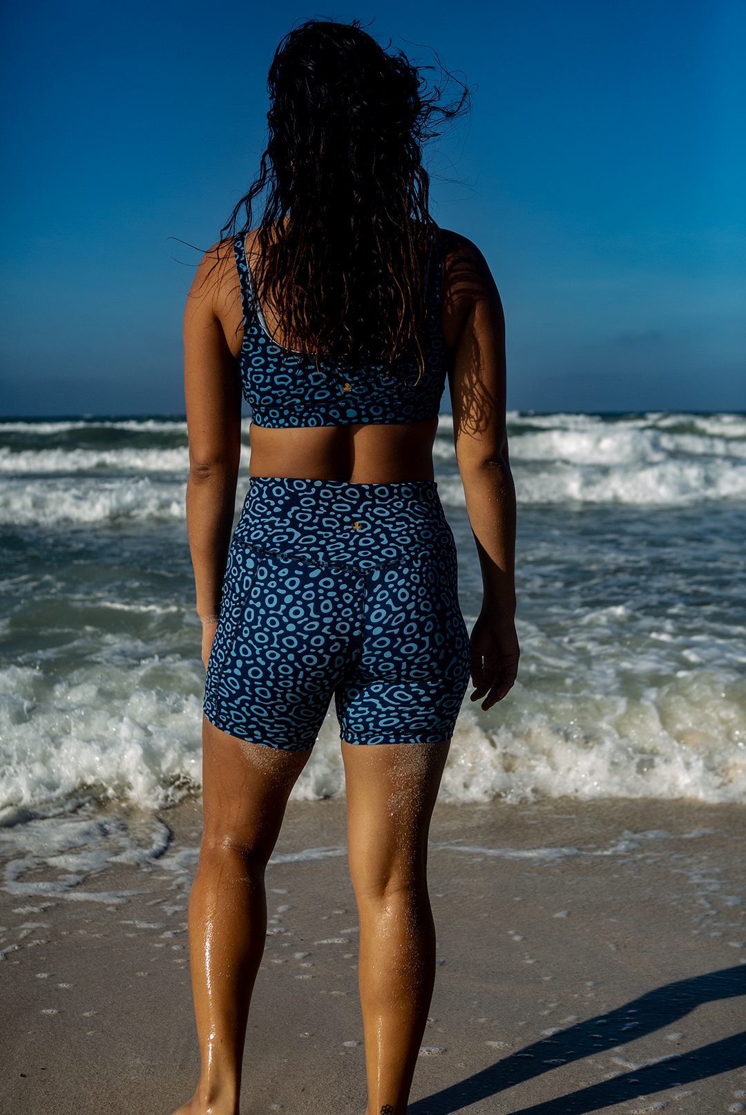 A woman wears NAUTRA’s Eagle Ray Print Biker Shorts on the coast. These high-waist workout shorts feature a side pocket, blending performance with ocean-inspired design.
