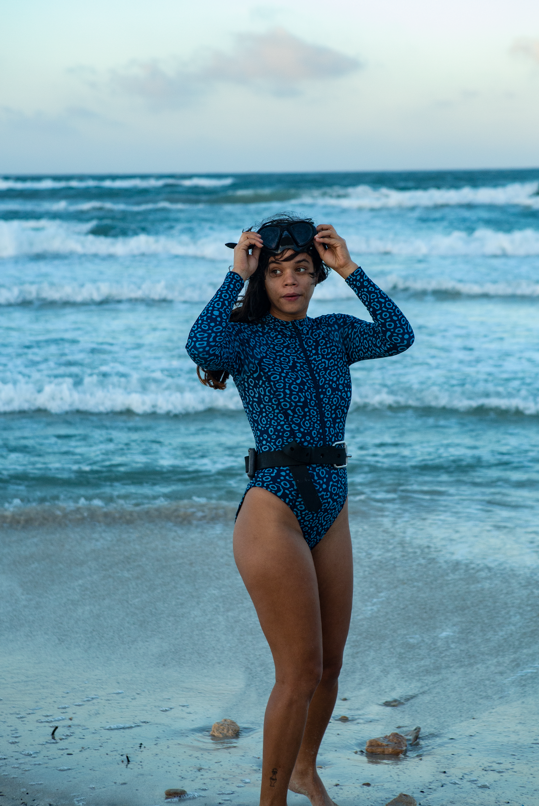 A woman wears NAUTRA’s Women&#39;s Long-Sleeve One-Piece Swimsuit in Eagle Ray Print on a sandy beach, with waves crashing behind her. This eco-friendly surf swimsuit is designed for adventure.