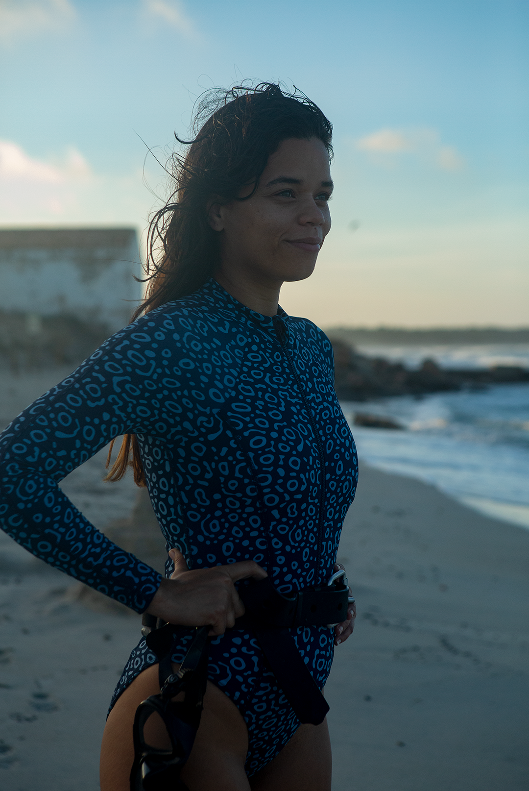 A woman wears NAUTRA’s Women's Long-Sleeve One-Piece Swimsuit in Eagle Ray Print on a sandy beach, with waves crashing behind her. This eco-friendly surf swimsuit is designed for adventure.