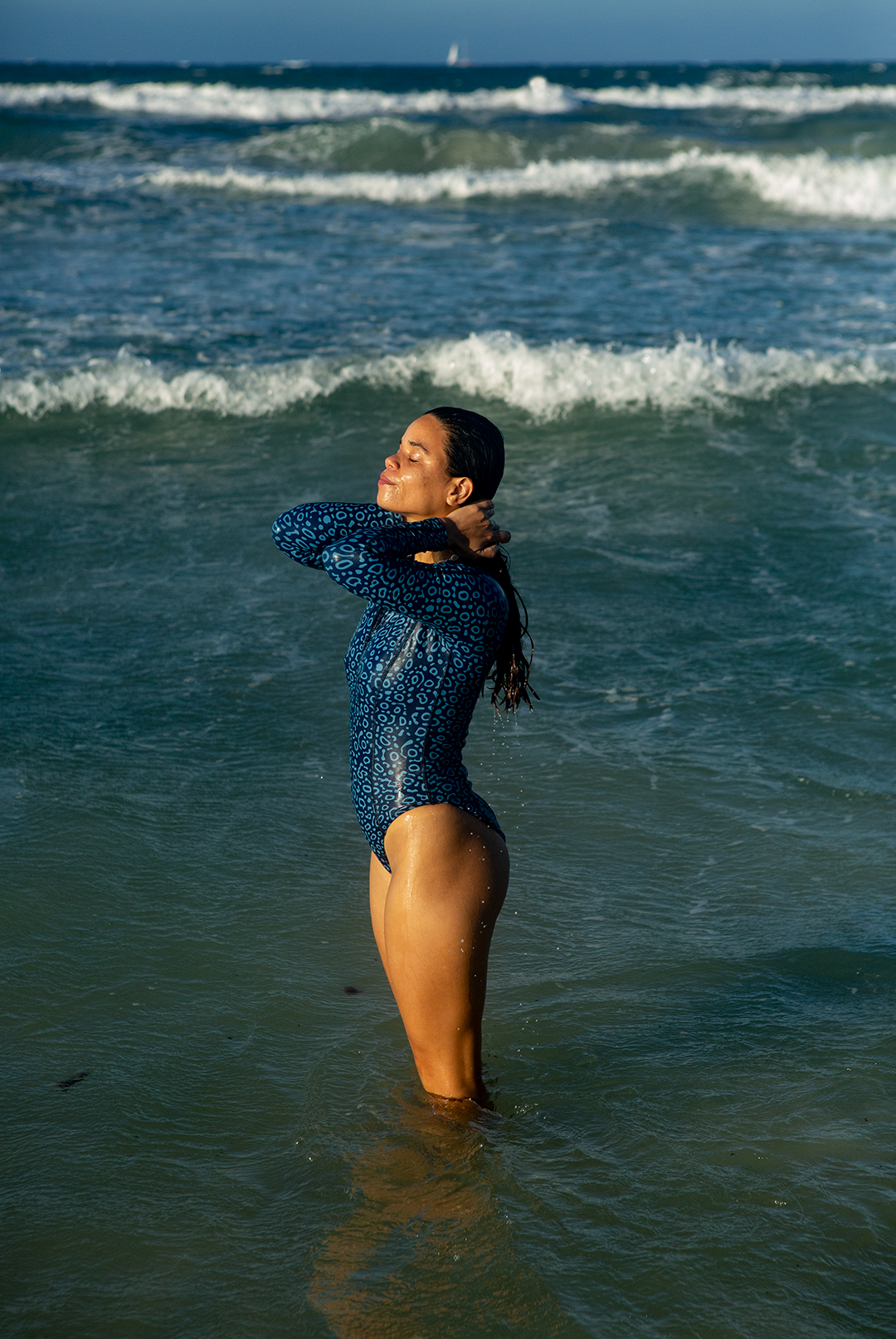 A woman wears NAUTRA’s Women&#39;s Long-Sleeve One-Piece Swimsuit in Eagle Ray Print on a sandy beach, with waves crashing behind her. This eco-friendly surf swimsuit is designed for adventure.
