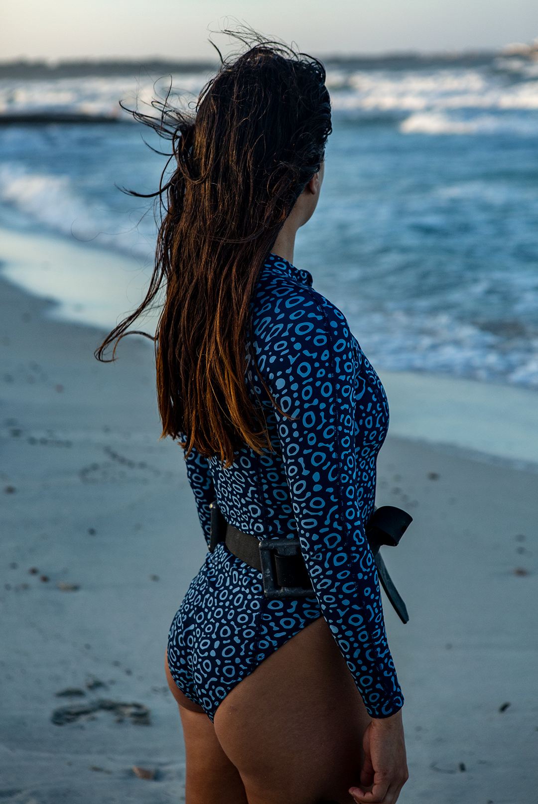 A woman wears NAUTRA’s Women's Long-Sleeve One-Piece Swimsuit in Eagle Ray Print on a sandy beach, with waves crashing behind her. This eco-friendly surf swimsuit is designed for adventure.