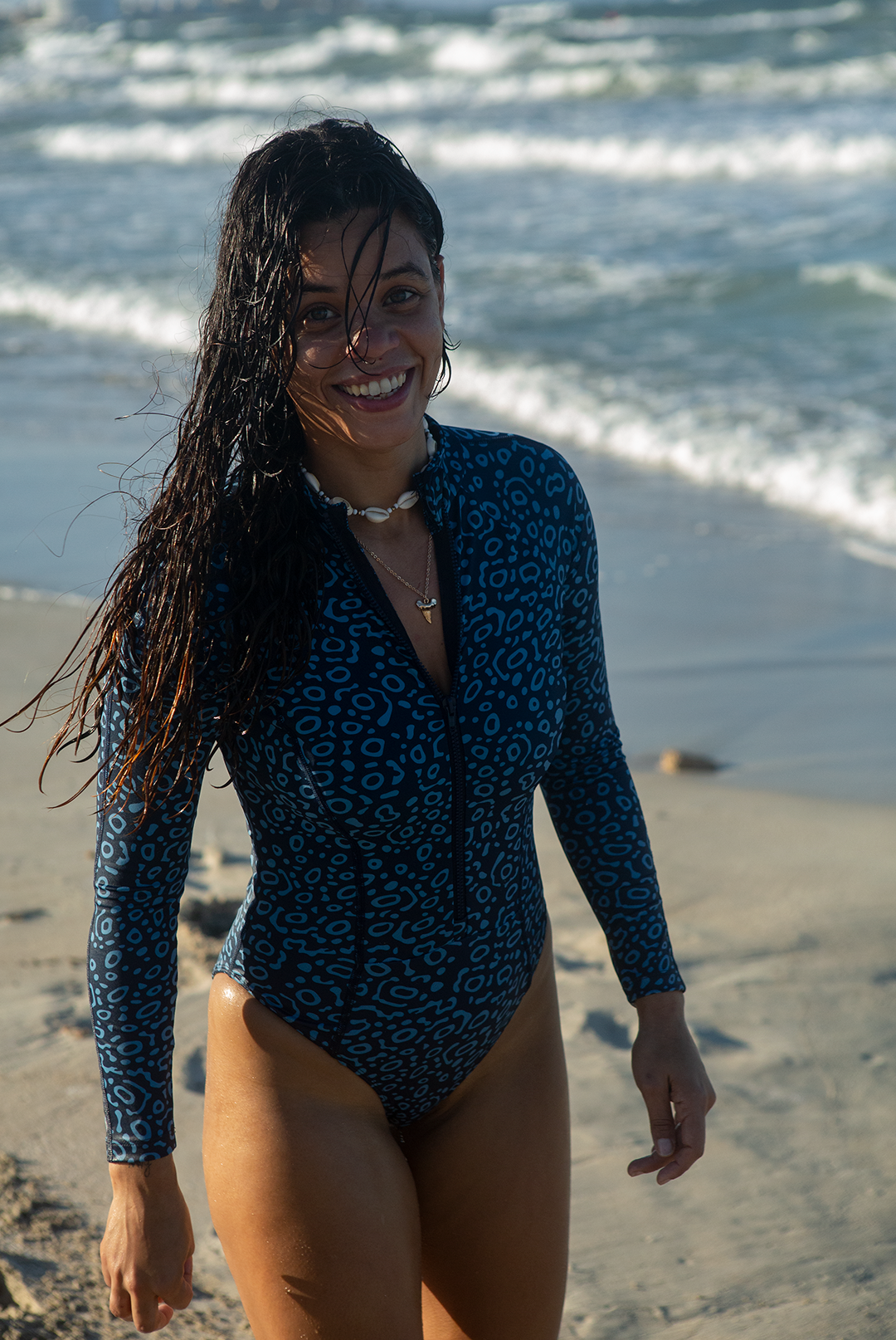A woman wears NAUTRA’s Women's Long-Sleeve One-Piece Swimsuit in Eagle Ray Print on a sandy beach, with waves crashing behind her. This eco-friendly surf swimsuit is designed for adventure.