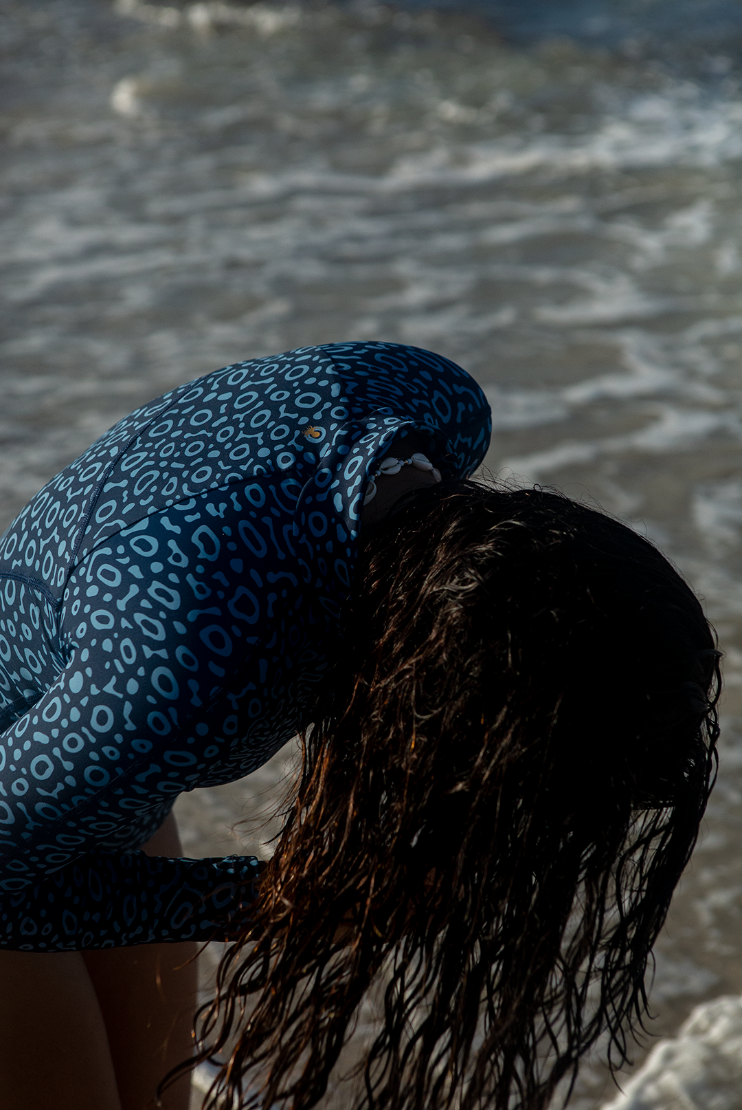 A woman wears NAUTRA’s Women's Long-Sleeve One-Piece Swimsuit in Eagle Ray Print on a sandy beach, with waves crashing behind her. This eco-friendly surf swimsuit is designed for adventure.