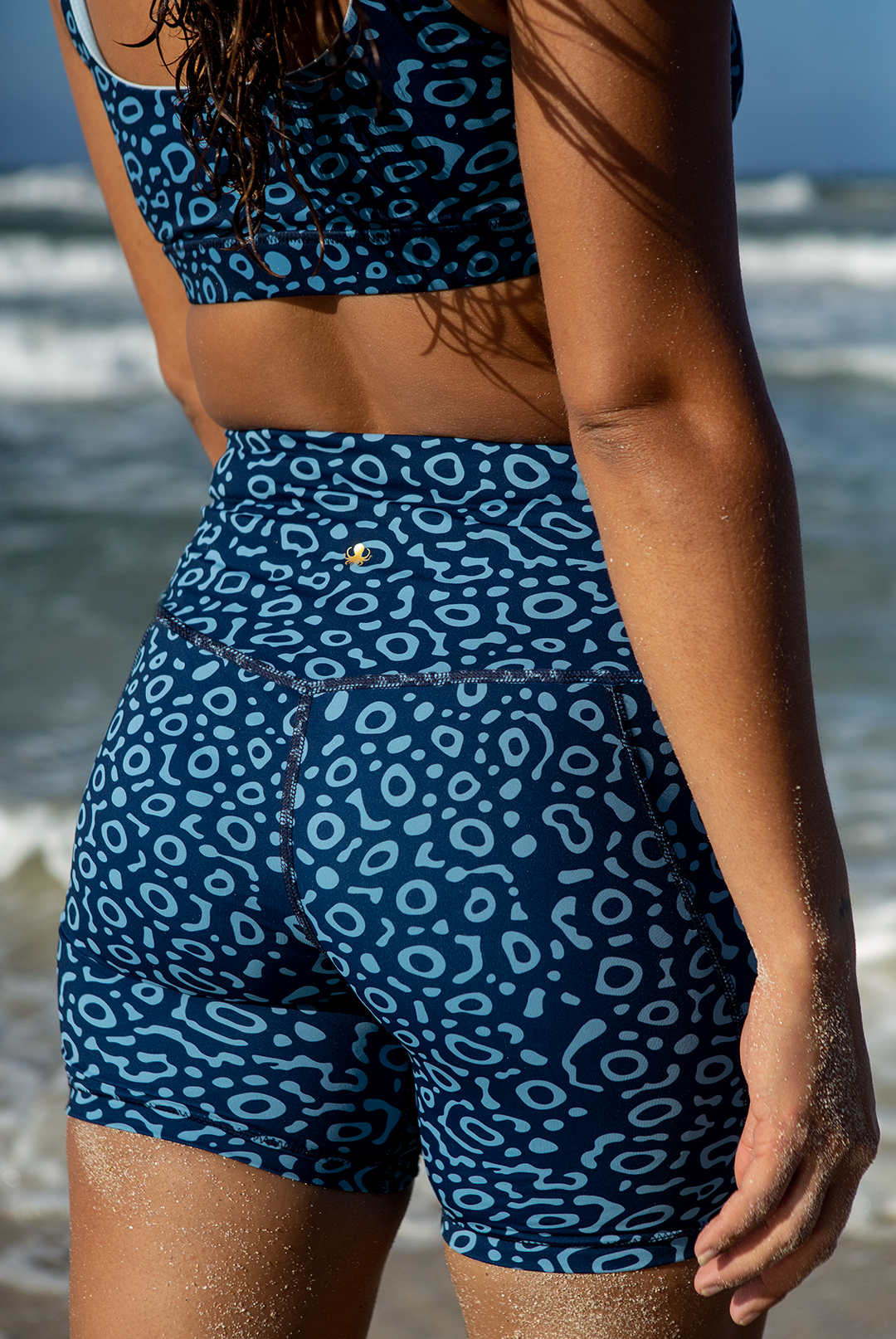 A woman wears NAUTRA’s Eagle Ray Print Biker Shorts on the coast. These high-waist workout shorts feature a side pocket, blending performance with ocean-inspired design.
