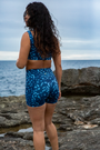 A woman wears NAUTRA’s Whale Shark Print Biker Shorts on a rugged shoreline. These high-waist workout shorts with a side pocket are built for movement and style.