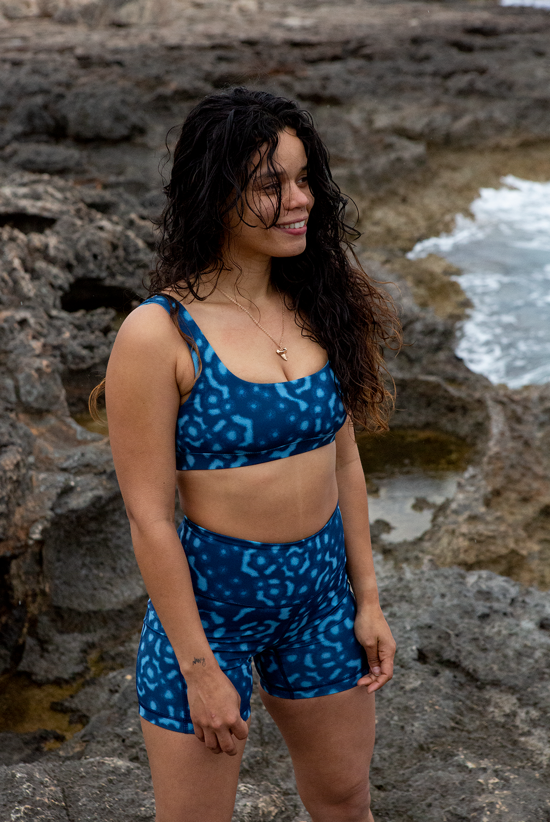 A woman wears NAUTRA’s Whale Shark Print Biker Shorts on a rugged shoreline. These high-waist workout shorts with a side pocket are built for movement and style.