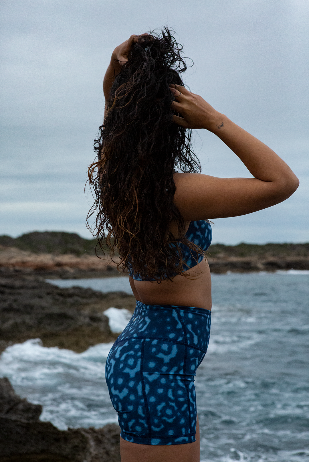 A woman wears NAUTRA’s Whale Shark Print Biker Shorts on a rugged shoreline. These high-waist workout shorts with a side pocket are built for movement and style.