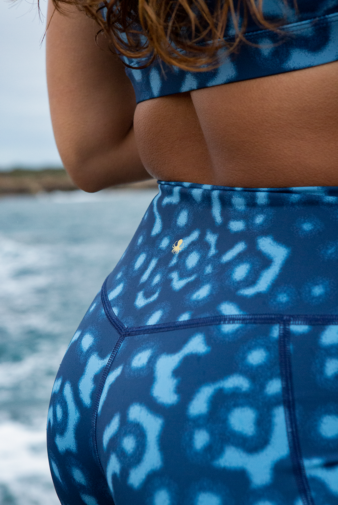 A woman wears NAUTRA’s Whale Shark Print Biker Shorts on a rugged shoreline. These high-waist workout shorts with a side pocket are built for movement and style.