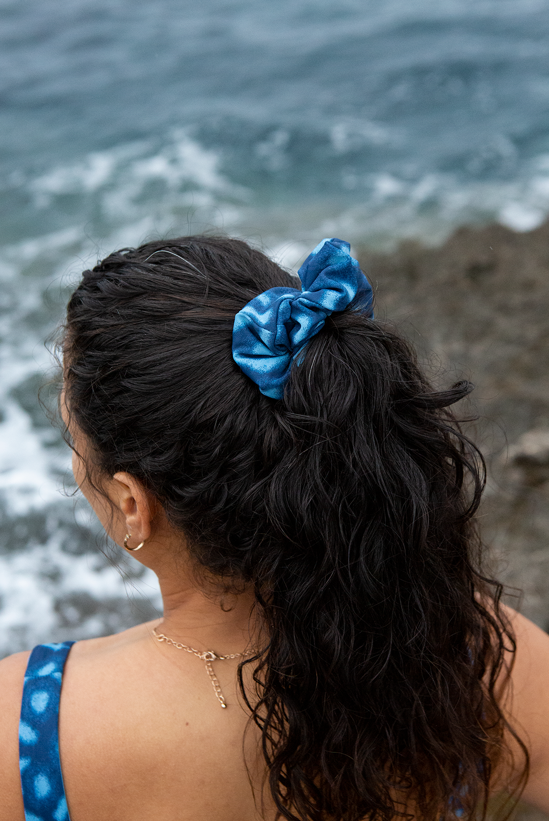Model standing on a rocky coastline with waves crashing in the background, wearing NAUTRA’s Whale Shark Print Scrunchie. Inspired by the unique patterns of whale sharks, this ocean-conscious accessory ties together style and sustainability.