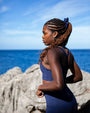 Model wearing NAUTRA Bluefin navy recycled strappy sports bra bikini top, back view, coastal ocean setting.