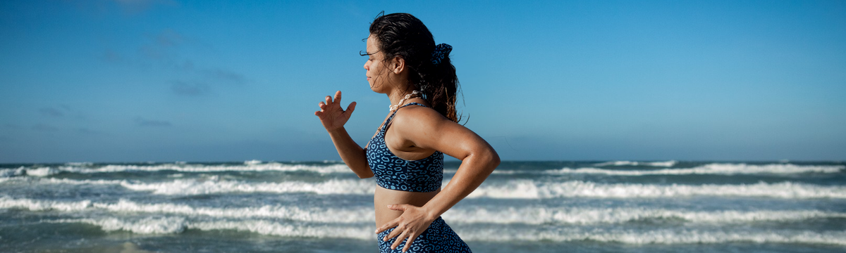 A woman in a NAUTRA eagle ray  patterned sports bra and matching bottoms stands on a beach with ocean waves and a clear blue sky in the background.