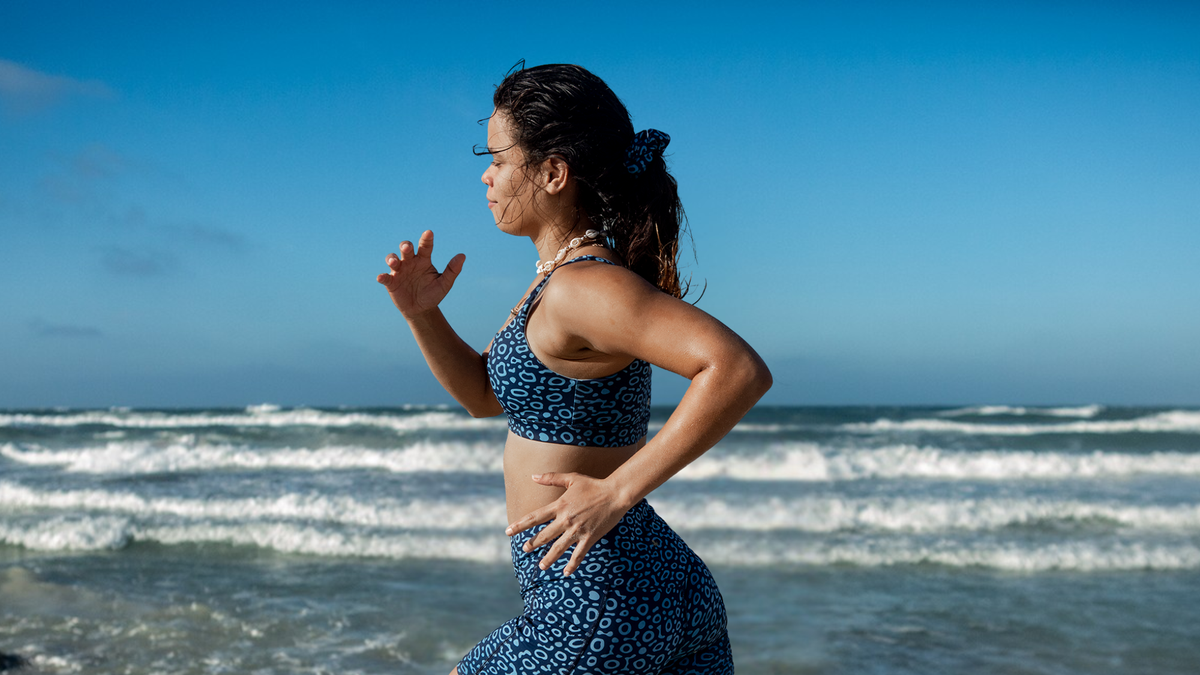 A woman in a NAUTRA eagle ray patterned sports bra and matching leggings runs along the shoreline of the ocean under a clear blue sky.