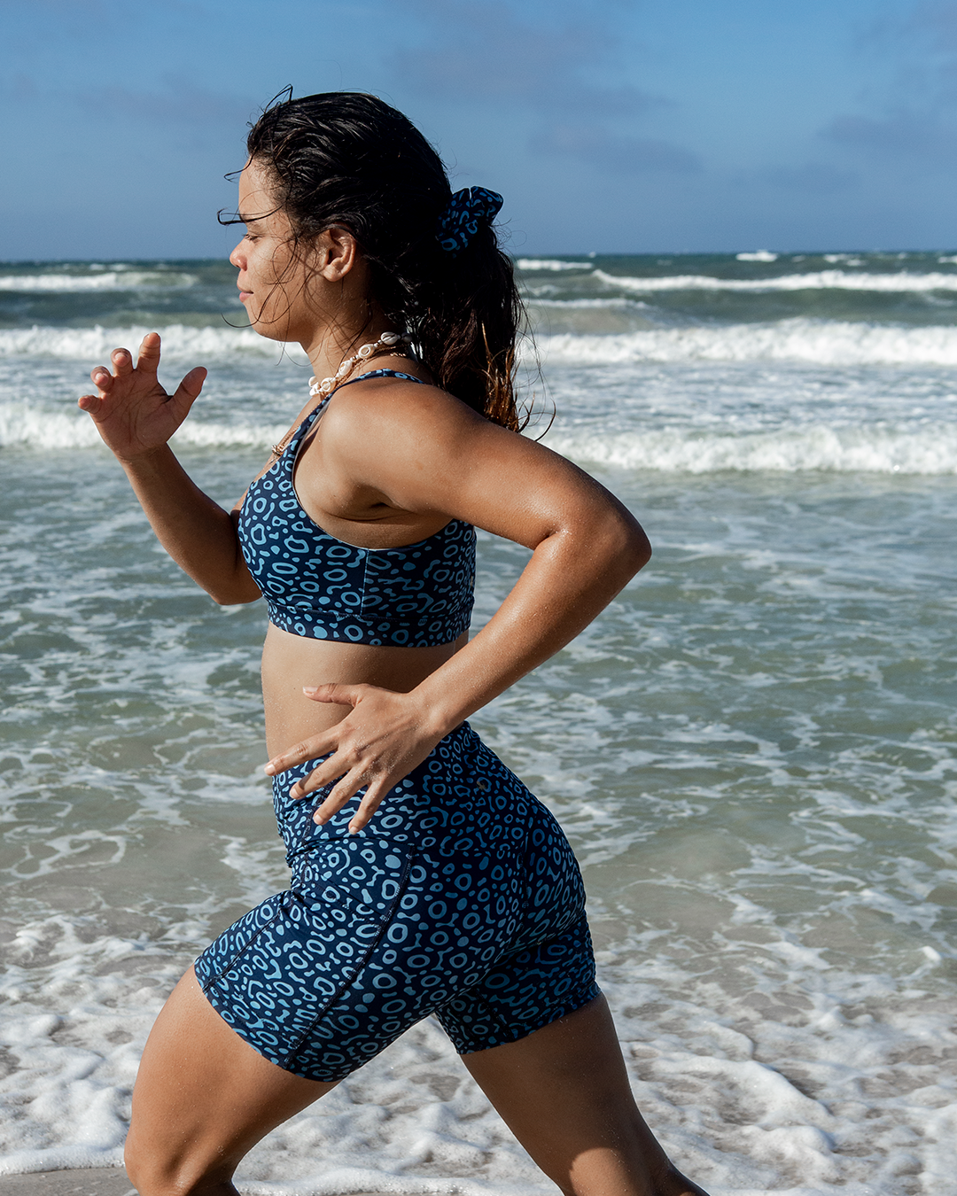 A woman running on a beach wearing a matching NAUTRA eagle ray patterned sports bra and biker shorts set.