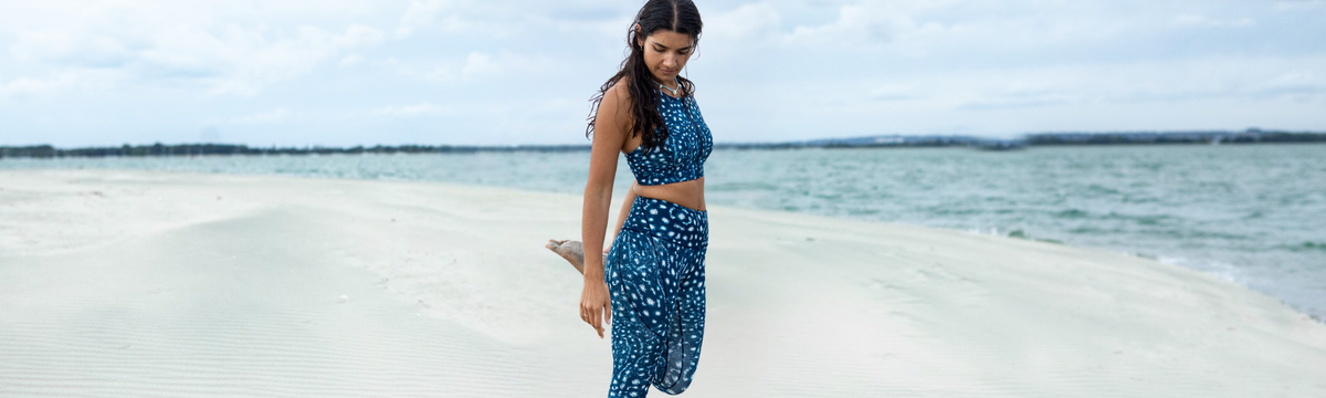 A woman stands on a sandy beach wearing a matching NAUTRA whale shark patterned two-piece outfit with a halter top and loose pants, with ocean and cloudy sky in the background.