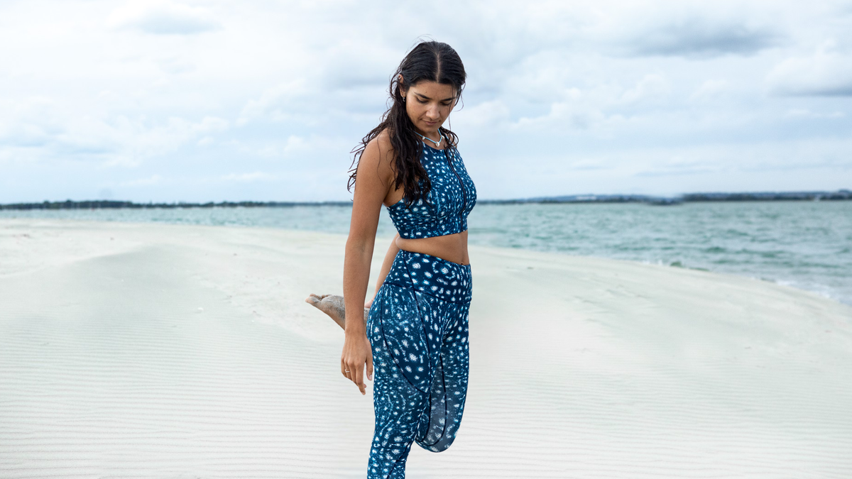 A woman stands on a sandy beach wearing a matching NAUTRA whale shark two-piece set with white polka dots, featuring a halter crop top and high-waisted pants.