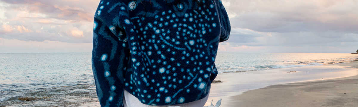 A person sitting on driftwood on a beach wearing the NAUTRA whale shark fleece, with the ocean and cloudy sky in the background.