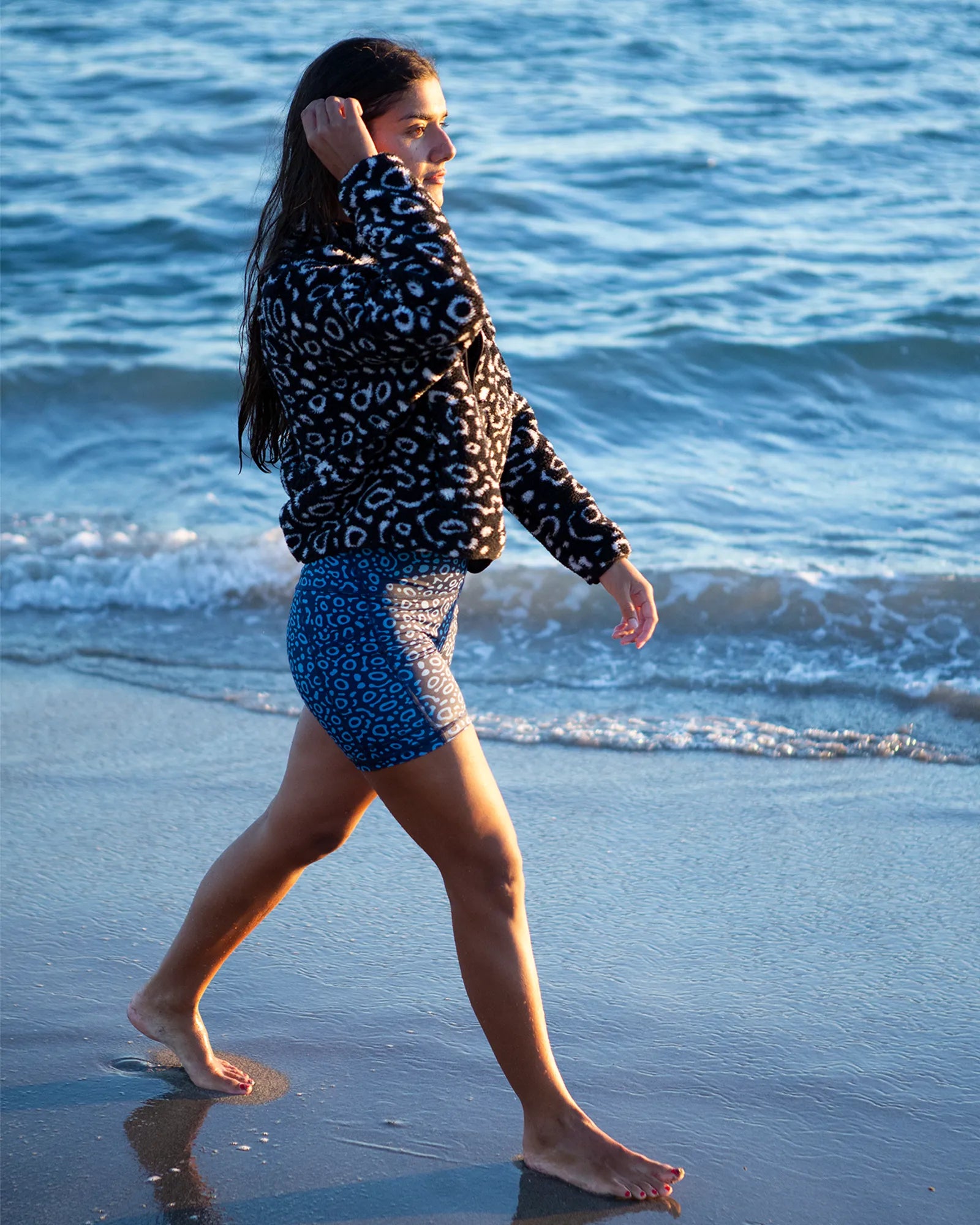 Model walking along the shoreline wearing NAUTRA Eagle Ray recycled high-waisted biker shorts and matching sherpa fleece, ocean in background.