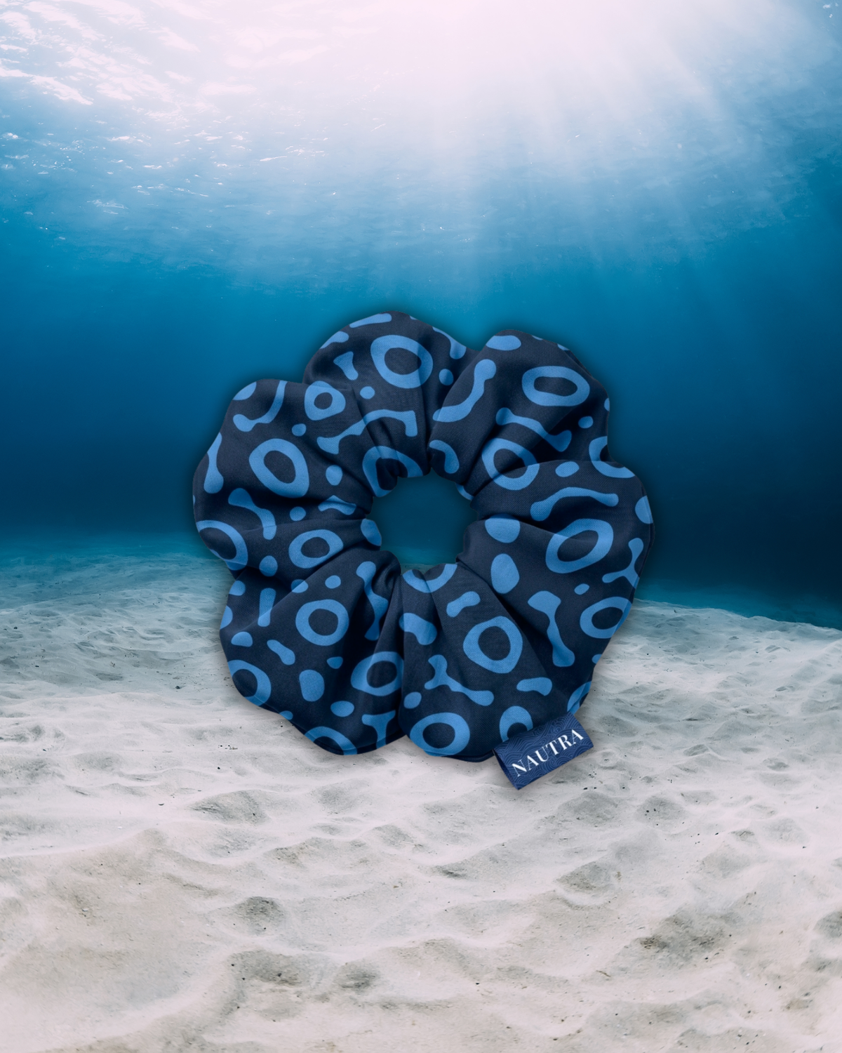 A navy blue scrunchie with a eagle ray  pattern, resting on a sandy ocean floor under clear blue water with sunbeams filtering down from the surface. A small tag on the scrunchie reads NAUTIA.