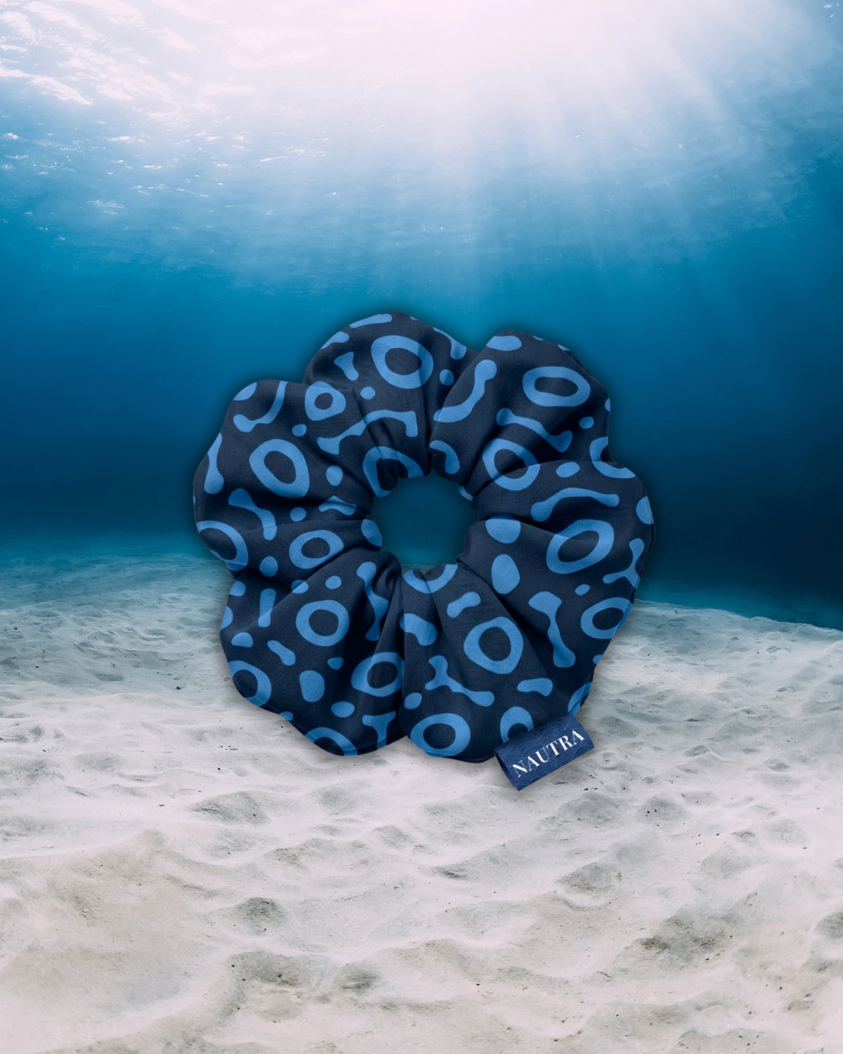NAUTRA eagle ray hair tie scrunchie, resting on a sandy ocean floor under clear blue water with sunbeams filtering down. A small tag on the scrunchie reads NAUTIA.
