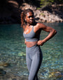 Model wearing NAUTRA Minke grey recycled high-waisted leggings beside clear coastal water, made from reclaimed ghost nets.