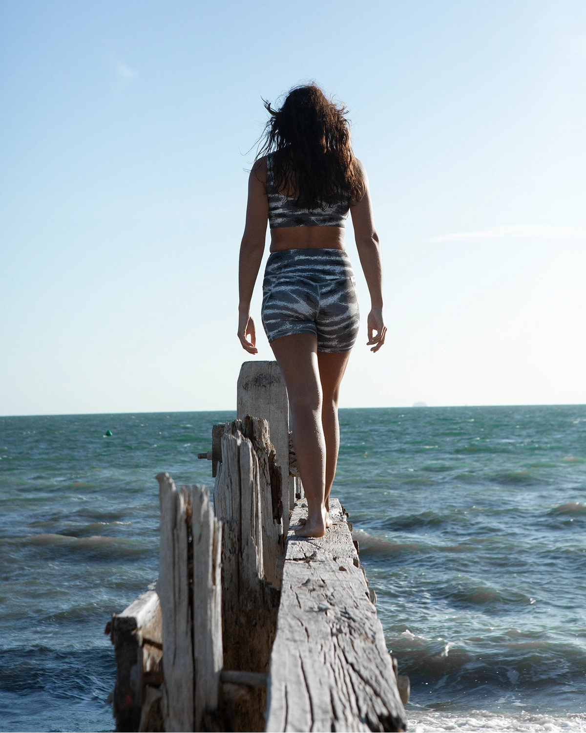 A woman seen from behind walking barefoot on a weathered wooden dock extending into the ocean, wearing a NAUTRA Tiger shark scoop neck sports bra bikini top and high waisted biker shorts set set against a clear blue sky.