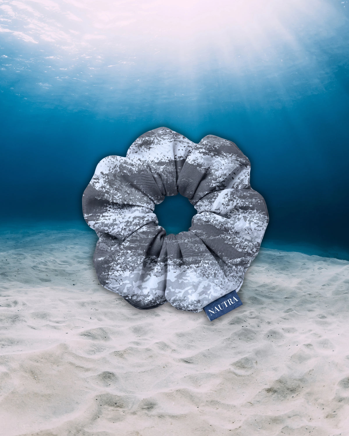 NAUTRA Tiger Shark hair tie scrunchie, resting on a sandy ocean floor under clear blue water with sun rays filtering down from the surface.