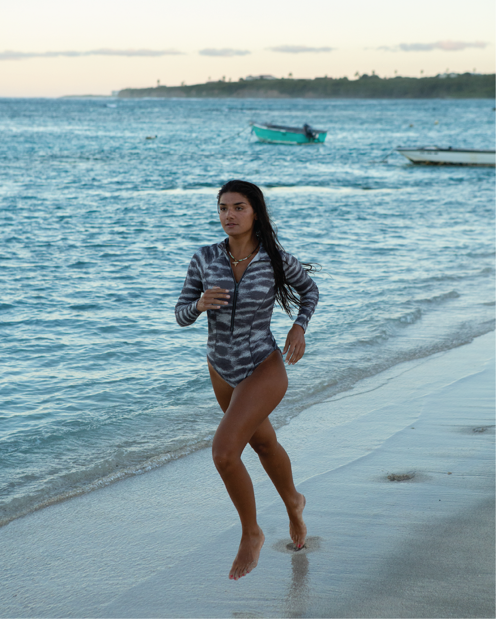 NAUTRA Tiger Shark long sleeve one-piece swimsuit in grey shark-inspired stripe print, worn running along the beach.