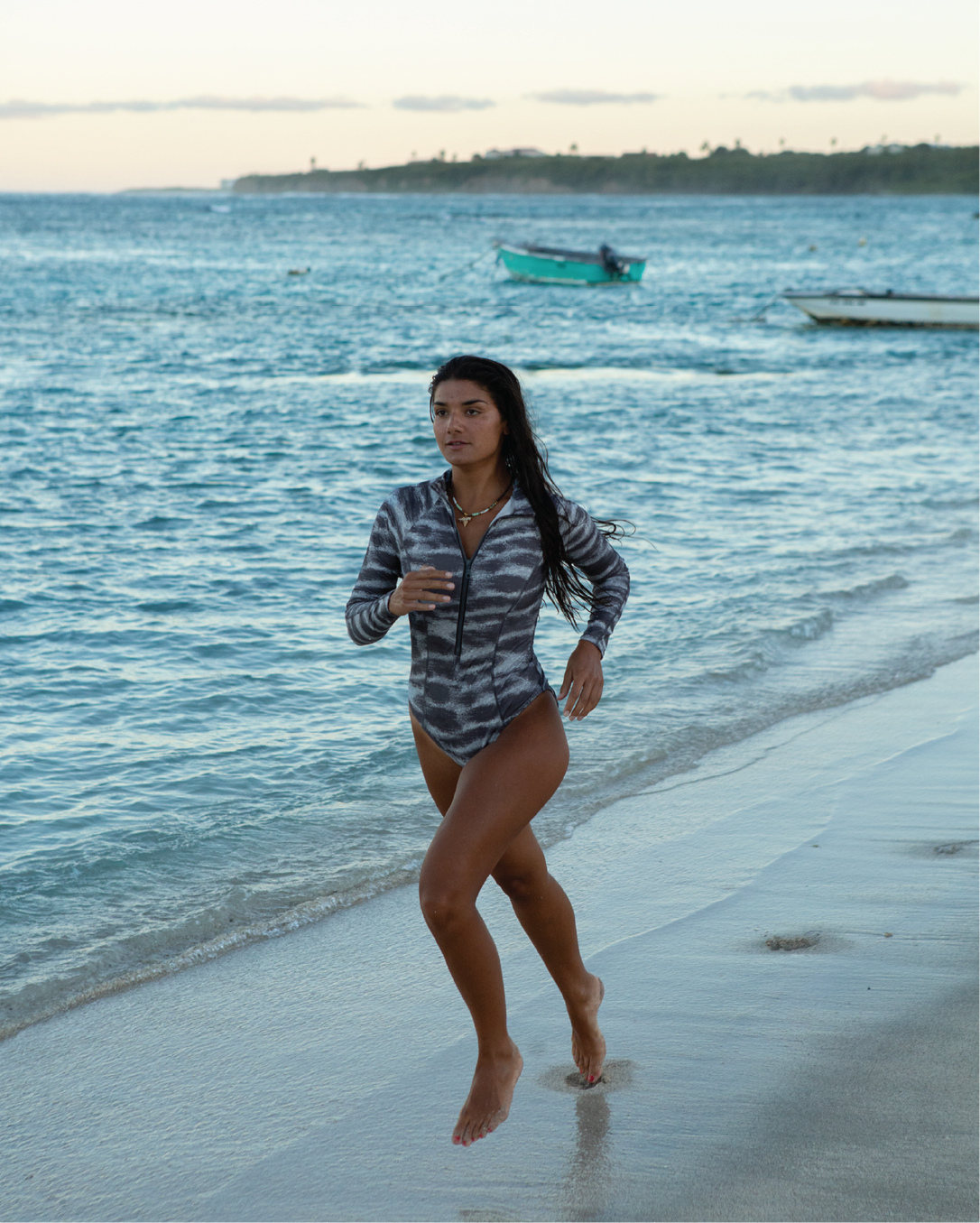 NAUTRA Tiger Shark long sleeve one-piece swimsuit in grey shark-inspired stripe print, worn running along the beach.