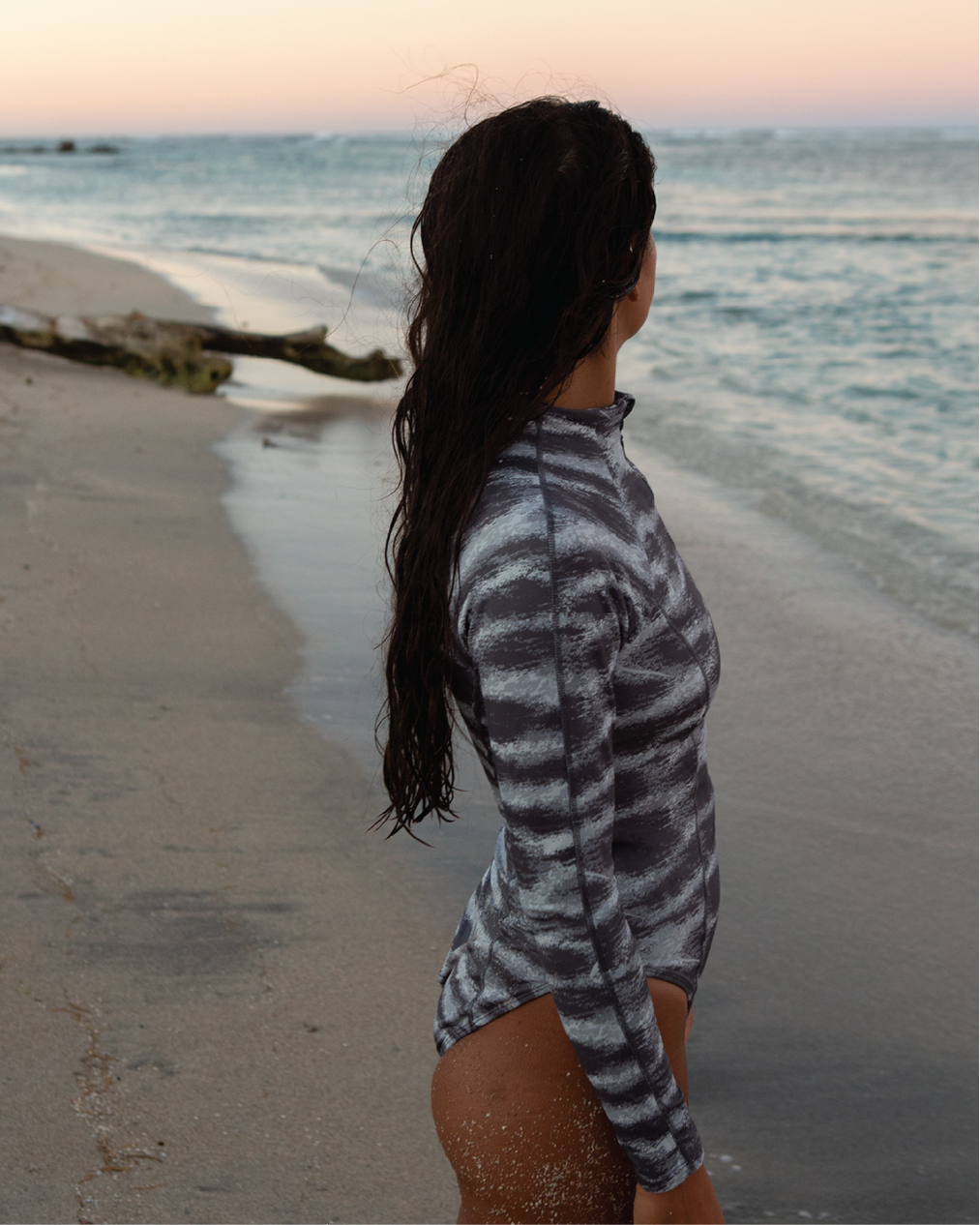 NAUTRA Tiger Shark long sleeve one-piece swimsuit with grey ocean-striped print, styled at sunset by the sea.