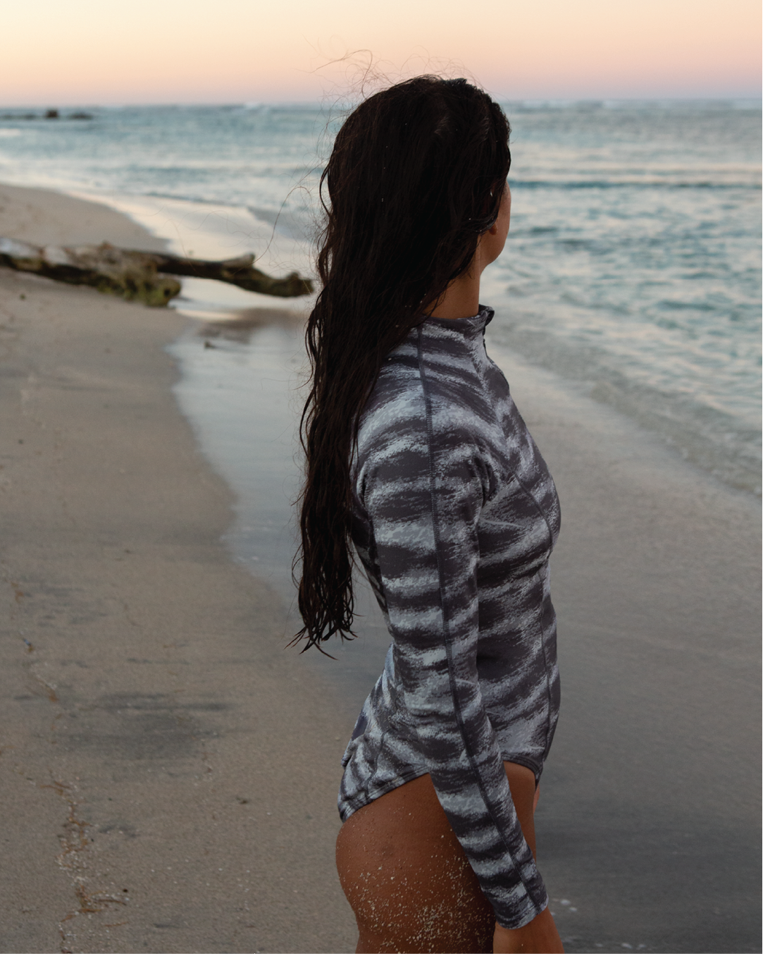 NAUTRA Tiger Shark long sleeve one-piece swimsuit with grey ocean-striped print, styled at sunset by the sea.