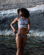A woman with dark curly hair stands in shallow water wearing a NAUTRA Tiger Shark Racer Back Sports bra bikini top and tie side bikini bottoms, with a rocky cliff background.