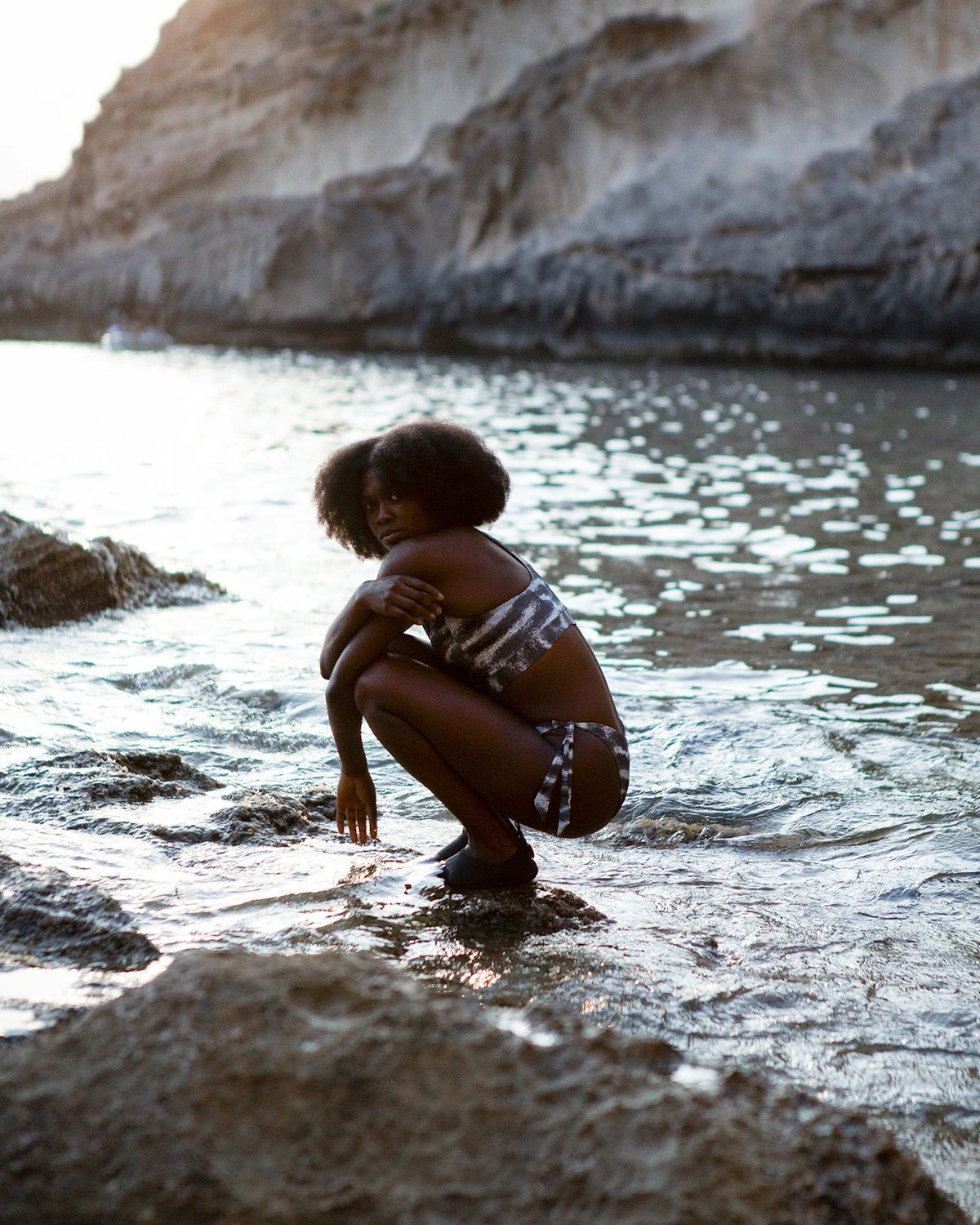 A woman crouching in shallow water at a rocky beach, wearing a NAUTRA Tiger Shark Racer Back Sports bra bikini top and tie side bikini bottoms. Cliffs rise in the background under warm sunlight.