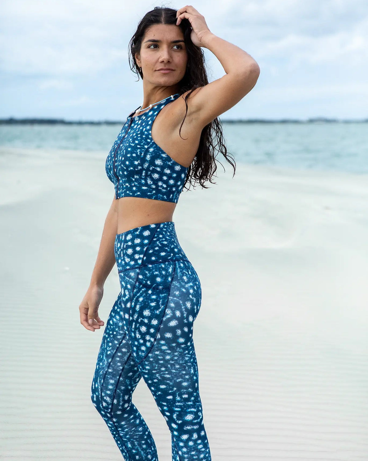 A woman stands on a sandy beach wearing NAUTRA's whale shark print set consisting of a sports bra and high-waisted leggings.