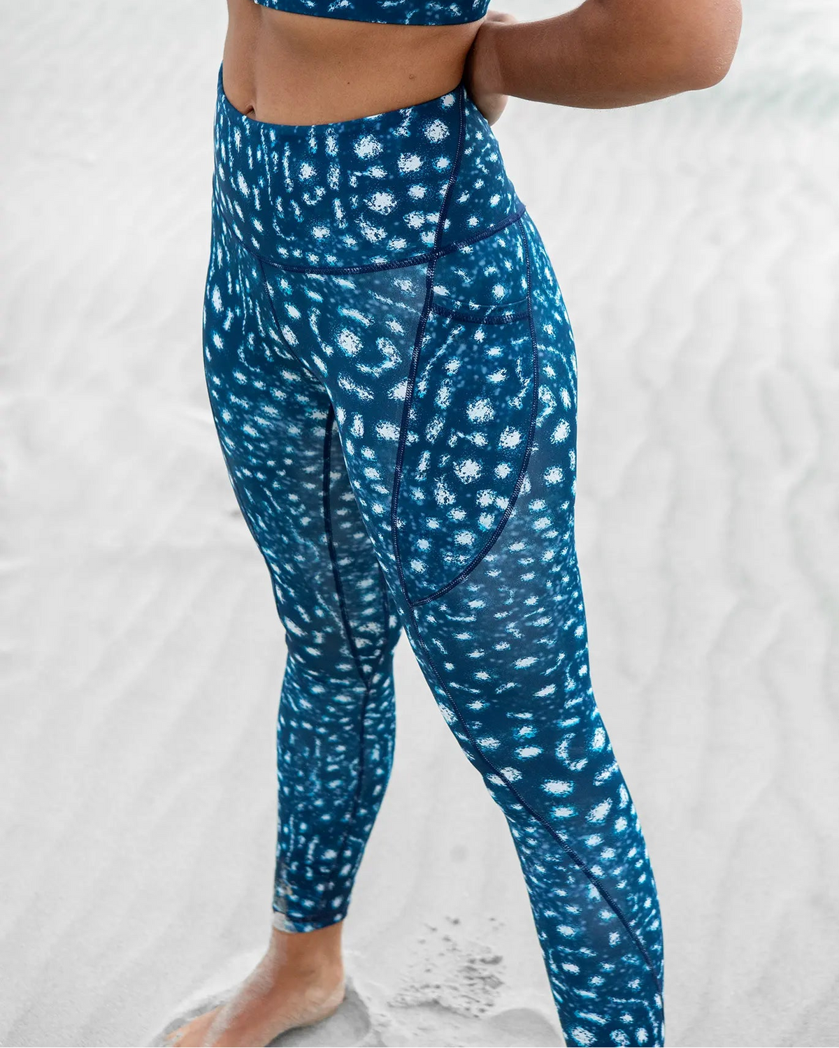 A woman standing on a sandy beach wearing NAUTRA's whale shark print set consisting of a sports bra and high-waisted leggings.